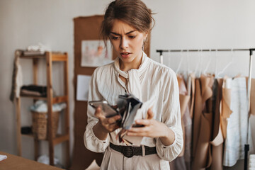 Thoughtful lady in stylish blouse holds phone and fabric samples. Young dark-haired woman in white...
