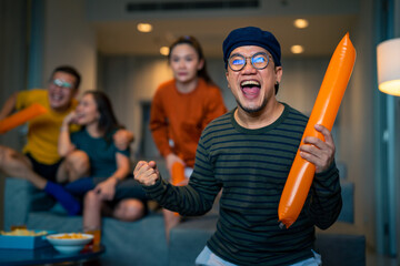 Group of Asian people friends sit on sofa watching and cheering sports games competition on TV...