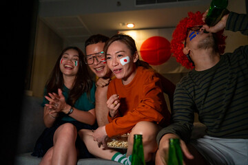 Group of Asian people friends sit on sofa watching and cheering sports games competition on TV together at home. Excited man and woman sport fans celebrate sport team victory in national sports match