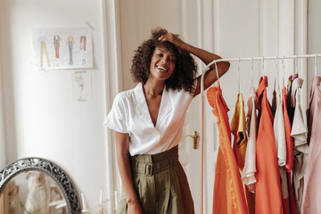 Happy excited dark-skinned curly woman in trendy khaki shorts and white blouse smiles widely, leans on hanger in white cozy room.