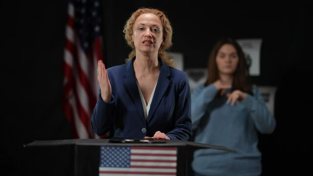 Blurred sign language translator showing nuts gesture and facepalm leaving as politician woman talking looking at camera. Interpreter walking away as dishonest lady presenting speech
