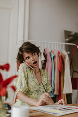 Surprised brunette curly short-haired woman in v-neck dress looks up, talks on phone and poses in office of fashion designer.