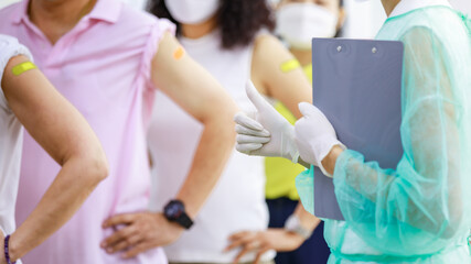 Nurse hand in full protection uniform and rubber gloves standing holding information board show thumb up to group of patients in vaccinated orderly straight line in hospital