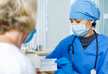 Doctor in blue hospital uniform face mask and rubber gloves with stethoscope send and explain Covid-19 vaccination record card certificate to Caucasian female patient in blurred foreground