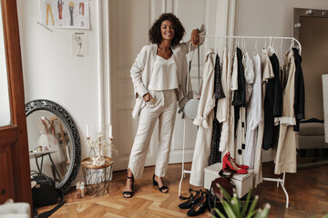 Pretty curly dark-skinned woman in trendy white pants, oversized jacket and stylish top looks into camera and leans in hanger in dressing room.
