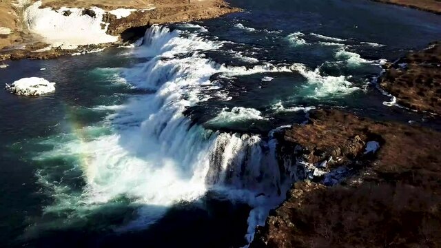An aeriel shot of rainbow cast over Faxi waterfall from a river that cuts through the snow covered countryside in Iceland