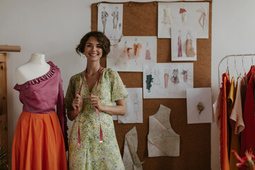 Attractive brunette short-haired woman in floral yellow dress smiling sincerely, holding measuring tape and posing near mannequin.