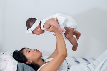 Lifestyle portrait of mother and daughter in white dress. Mother laying down on bed, lifting up her baby high. 
