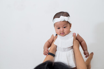 Young mother with her baby girl on bed playing happily. Southeast asian family, mother and daughter wearing white fashionable dress. Mom lifting up baby cheerfully. 