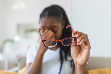 African girl in glasses rubs her eyes, suffering from tired eyes, ocular diseases concept. young woman holds glasses with diopter lenses and rubs her eyes from fatigue fatigue of vision