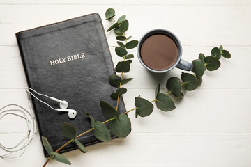 Holy Bible, cup of coffee, earphones and eucalyptus branch on light wooden background