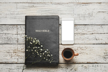 Holy Bible, cup of coffee, mobile phone and flowers on light wooden background