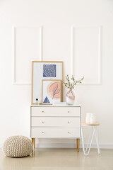 Vase with blossoming branches and pictures on chest of drawers near light wall