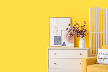 Vase with blossoming branches on chest of drawers near color wall