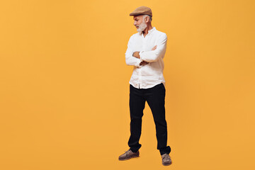 Adult man in cap, suit poses on orange background. Bearded handsome guy in white long-sleeved shirt and black modern pants