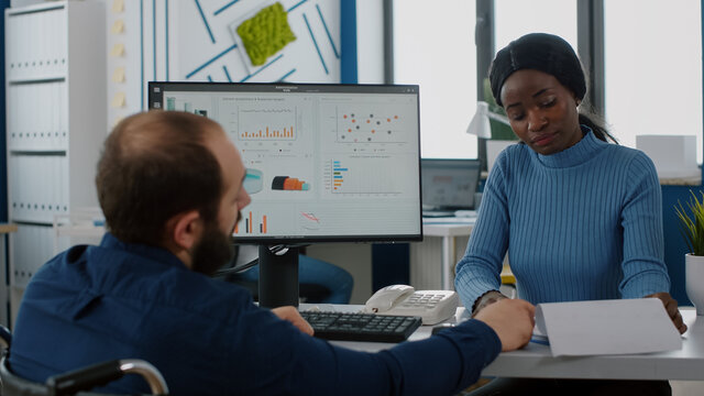 Handicapped businessman and african employee working at financial project discussing in start up company, checking statistic info looking at pc showing data prosessing on display for regarding economy