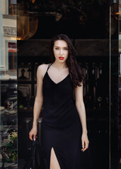 Fototapeta na wymiar Gorgeous young model woman with perfect dark hair looking at camera posing in the city wearing black evening dress. Beautiful sexy lady.