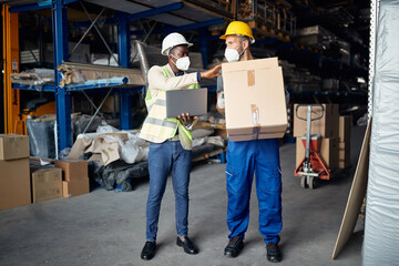 Black warehouse foreman uses laptop and checks packages carried by the worker at storage compartment.