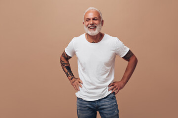 Tattooed man in good mood posing on beige background. Gray-haired guy in white T-shirt and blue...