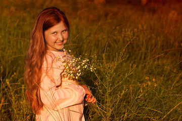   Portrait of cute smiling caucasian girl walking on the meadow and holding a bouquet of wildflowers.  Summer sunset.  Happy Childhood concept. Nature background. Copy space.