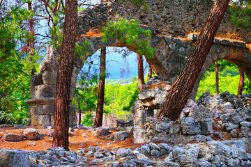 Ruins of Roman aqueduct and straggling heaps of stones in ancient city of Phaselis and beautiful turkish nature - pine trunks, bright green foliage and blue stormy sky. Antalya province, Turkey.