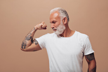 Adult man in white shirt demonstrates his muscles. Gray-haired guy with tattoos on his hands in...