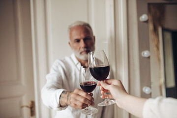 White haired man clinking wine glass. Bearded gray-haired guy with pensive face holds red wine in his hand in bright apartment