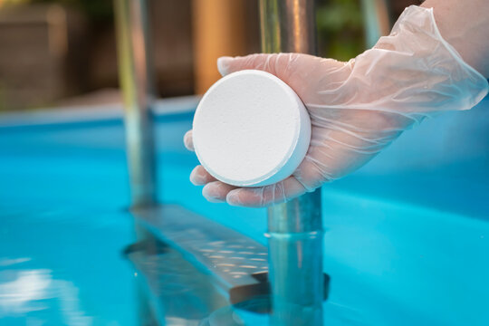 Human hand in protective glove holding white pill on background of blue clear water of swimming pool close up. Concept of maintenance, disinfection of water in pool.