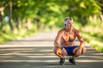 Muscular and fit older man squats as he rests after doing sport outdoors in the nature