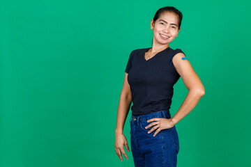 Middle aged Asian woman smiling showing her arm with bandage patch showing she got vaccinated for Covid 19 virus on green background. Concept for Covid 19 vaccination