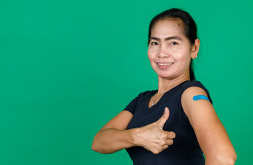 Asian middle aged woman smiling and pointing at her arm with bandage patch showing she got vaccinated for Covid 19 virus on green background. Concept for Covid 19 vaccination