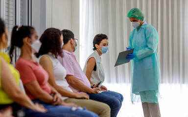Young female citizen wears face mask in Coronavirus vaccinating queue line sitting checking her personal information from paper board in hand of full protective suit nurse who standing nearby