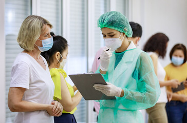 Obraz premium Young female nurse in full hazard protection uniform holding paper board asking personal information from senior caucasian woman patient while others wait in queue line in blurred background
