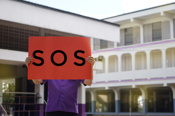 Text " SOS" on paper sign held by laborer standing in front of the factory. Concept : emergency  asking for help. Blurred background and light.      