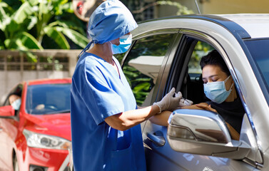 Female doctor in blue hospital uniform and face mask stand near car holding syringe in hands wears rubber gloves injecting covid 19 vaccine to drive through patient shoulder on vaccination queue