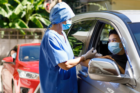 Female Doctor In Blue Hospital Uniform And Face Mask Stand Near Car Holding Syringe In Hands Wears Rubber Gloves Injecting Covid 19 Vaccine To Drive Through Patient Shoulder On Vaccination Queue