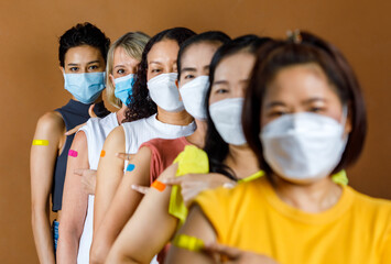 Diverse group of multinational ethnic female patients wears face mask stand in line by height order look at camera pointing colorful plasters on shoulder together after vaccinated - obrazy, fototapety, plakaty