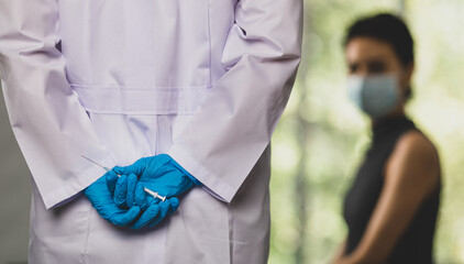 Doctor in white lab coat and blue rubber gloves hold and hiding vaccine syringe needle in hand behind back before walking to female patient who sitting and waiting frightened in blurred background