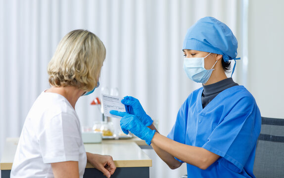 Young female doctor in blue hospital uniform face mask and rubber gloves sit showing explaining about Covid-19 vaccination record card certify to blonde Caucasian patient - Powered by Adobe