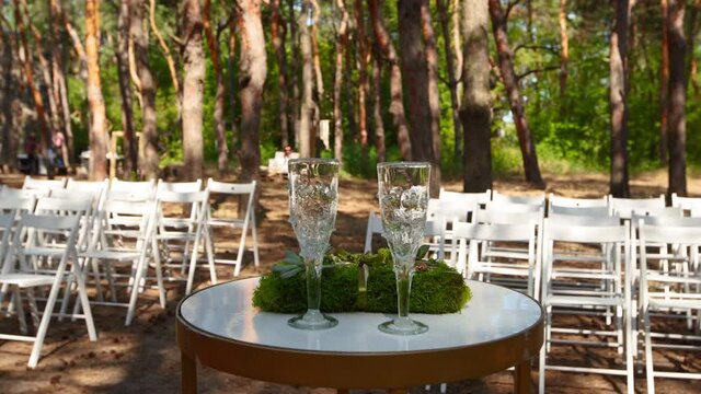 Wine glasses, wedding rings bedding of grass and moss on white table next to bohemian tipi arch decorated in boho style with flowers and candles and white chairs for guests.