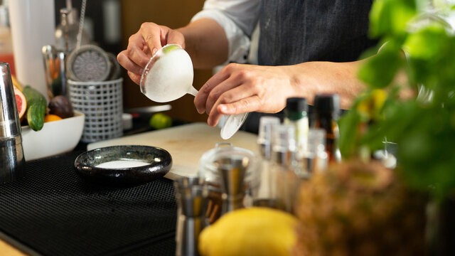 A Bartender Rimming The Frozen Cocktail Glass With a Lime Wedge. Surrounded With Bar Equipment and Cocktail Ingredients. Wearing a Dark Grey Apron With a White Shirt.