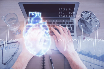 Double exposure of man's hands typing over computer keyboard and human heart hologram drawing. Top view. Medical education concept.