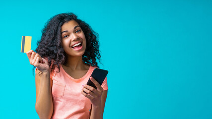 Fototapeta premium Happy smiling girl posing with credit card and mobile phone in hands, isolated over blue background