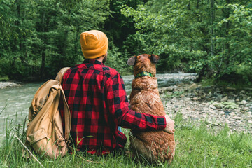 Back view of a young bearded hipster tourist man sitting together with his dog buddy at the river bank, relaxing after long hiking journey in the forest, enjoying great scenery - 445342459