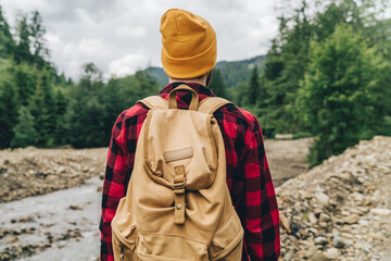 Young male tourist with a travel backpack standing near the river enjoying the scenery of evergreen forest and mountains