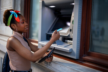 Fototapeta na wymiar Beautiful african women using ATM machine. Attractive young woman withdrawing money from credit card at ATM...