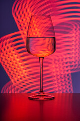 A crystal glass filled with pure water against a purple background and neon reddish streaks painted with light. Reflections, refractions, light backdrop, beverages and cocktails, lifestyle, abstract.