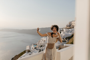 Cheerful brunette woman in stylish summer outfit shows tongue, v-sign and takes selfie in beautiful city with sea view.