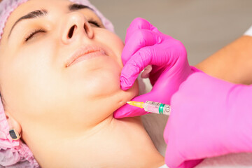 The cosmetologist makes lipolytic injection on the chin of a young woman against the double chin in a beauty salon - obrazy, fototapety, plakaty