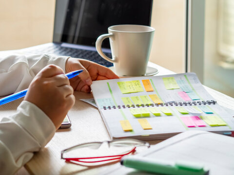 Close-up Of a businesswoman's hands organizing agenda with sticky notes at workplace. Color planning for productivity and remainder of important appointments. Desk with coffee cup, laptop and glasses.